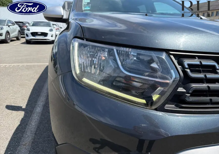 Gros plan sur le phare avant gauche et la calandre d’un Dacia Duster gris Comète métallisé, vue de face.