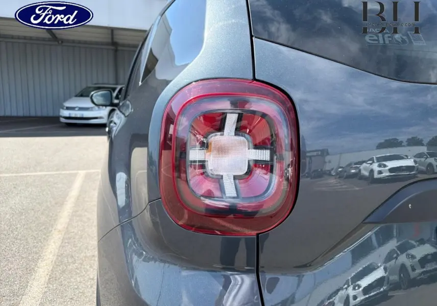 Gros plan sur le feu arrière droit du Dacia Duster gris Comète métallisé, avec reflet du ciel et voitures alentour.