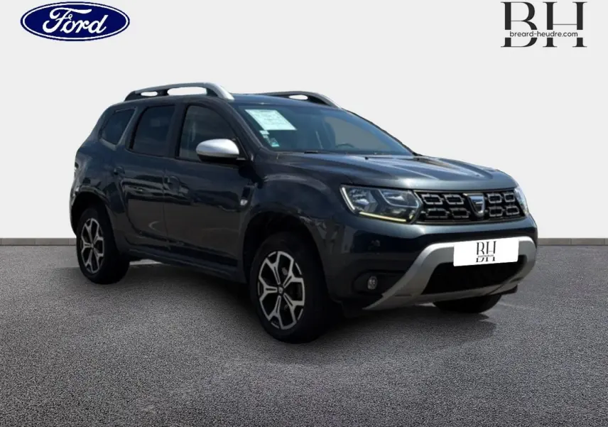 Dacia Duster gris Comète métallisé en 3/4 avant droit, avec barres de toit et jantes alliage visibles.