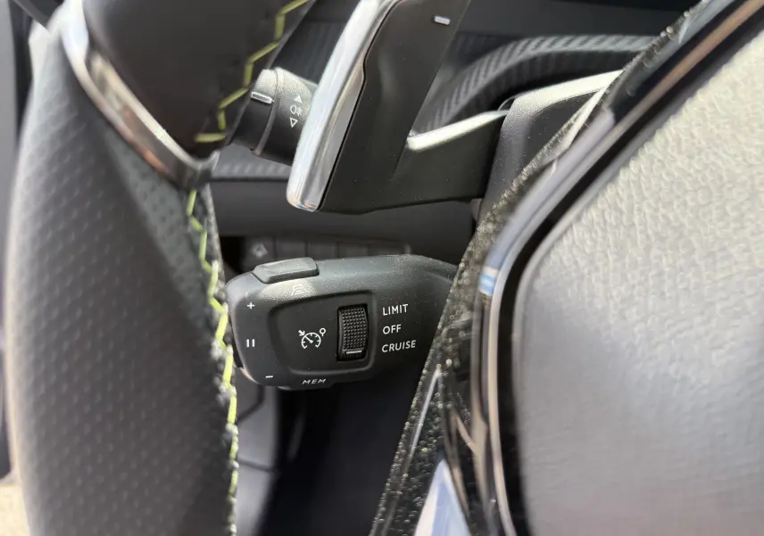 Gros plan sur la commande de régulateur de vitesse à gauche du volant noir perforé dans l'habitacle d'une Peugeot 208 gris Artense.