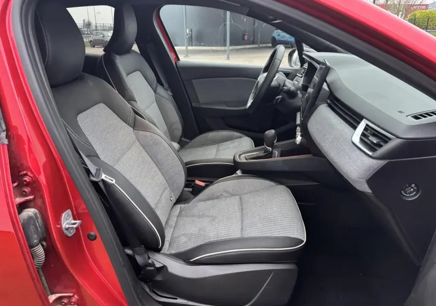 Vue intérieure côté conducteur de la Renault Clio V rouge flamme, montrant sièges gris mélangé et tableau de bord moderne.
