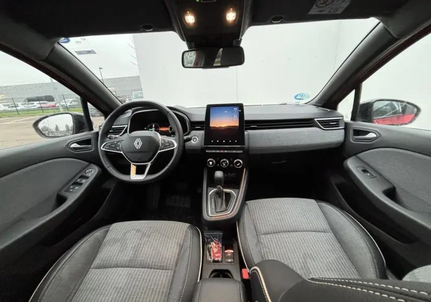 Vue intérieure avant de la Renault Clio V hybride, tableau de bord gris avec écran tactile vertical et volant multifonction.