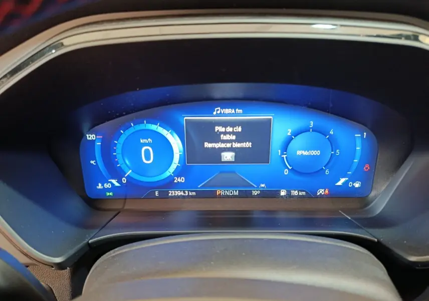 Tableau de bord numérique bleu du Ford Kuga 2024 affichant vitesse, compte-tours et message batterie clé faible.