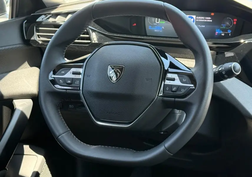 Vue rapprochée du volant multifonction noir de la Peugeot e-308 2025 avec tableau de bord numérique en arrière-plan.