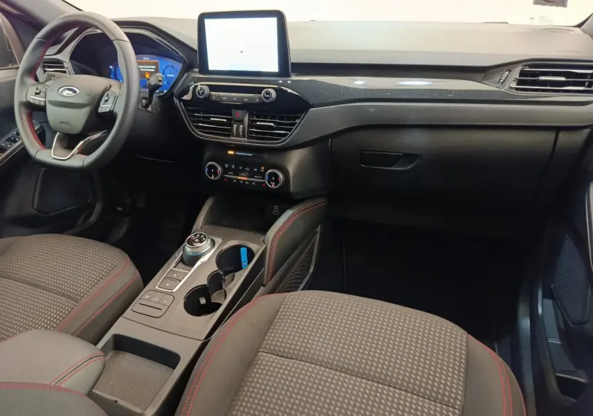 Vue intérieure avant droite du Ford Kuga 2024 St Line avec tableau de bord noir, écran tactile central et sièges tissu gris à surpiqûres rouges.