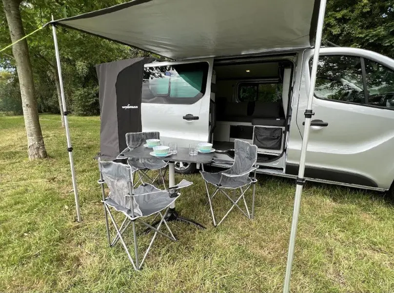 Vue latérale droite d’un Renault Trafic gris aménagé avec porte coulissante ouverte et auvent déployé sur un espace de pique-nique.