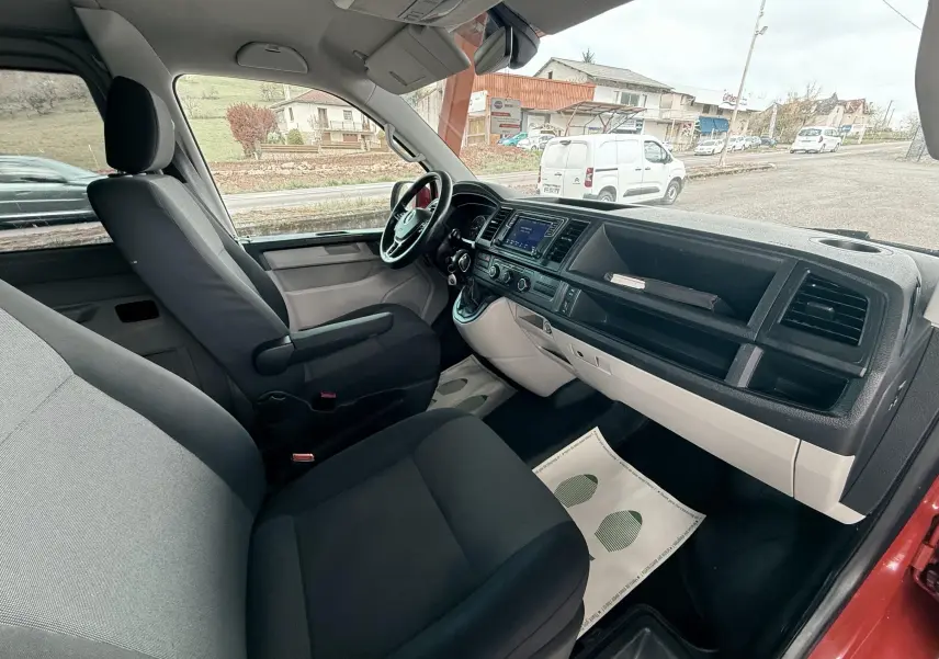Vue intérieure côté conducteur du Volkswagen Transporter rouge, montrant le tableau de bord et les sièges en tissu noir et gris.