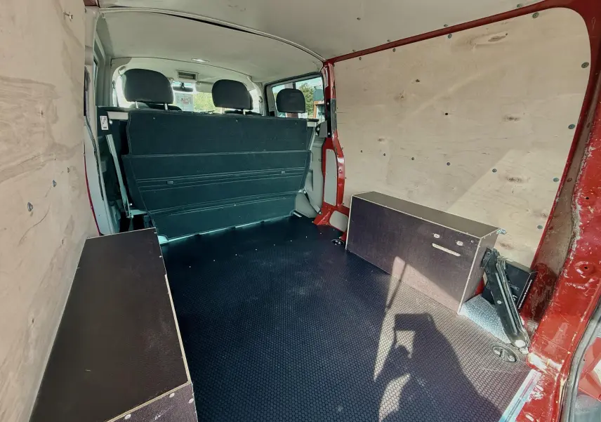 Vue intérieure arrière du Volkswagen Transporter rouge, montrant l’espace de chargement avec panneaux bois et banquette rabattue.