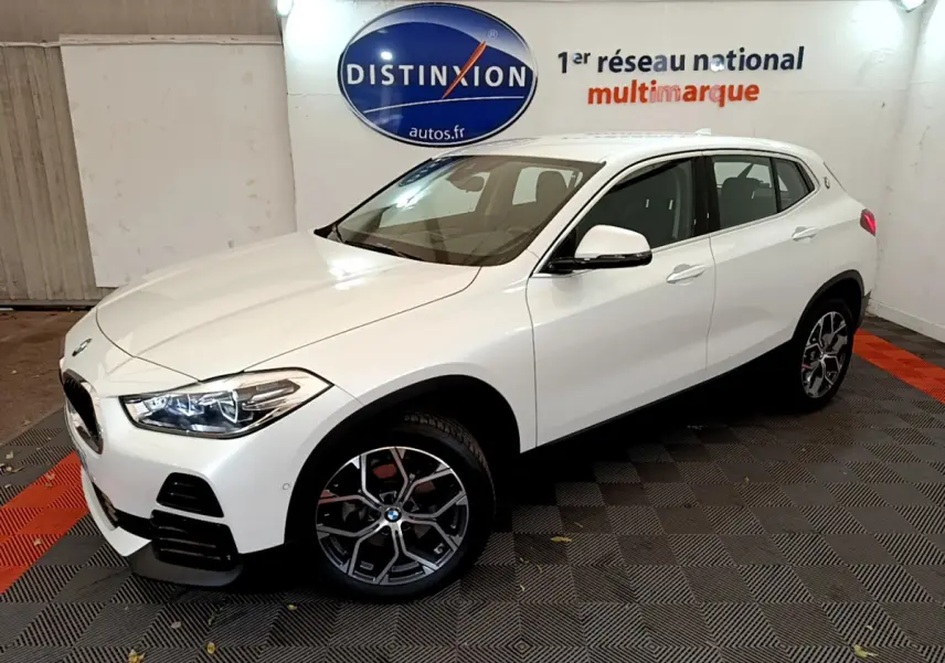 BMW X2 blanc en 3/4 avant droit dans un showroom avec jantes alliage et phares LED allumés.