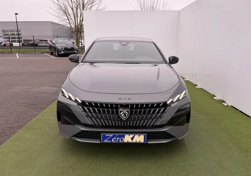 Vue frontale d'une Peugeot 308 III gris selenium avec calandre noire et logo lion lumineux sur fond de parking.