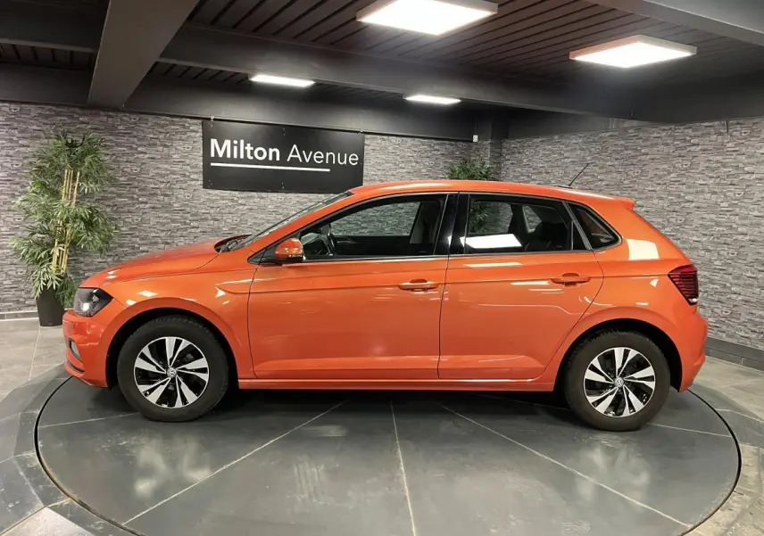 Volkswagen Polo orange vue de profil côté gauche, avec jantes alliage et intérieur visible en showroom.