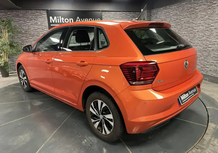 Vue 3/4 arrière droite d'une Volkswagen Polo orange métallisée avec jantes alliage et feux arrière LED dans un showroom.