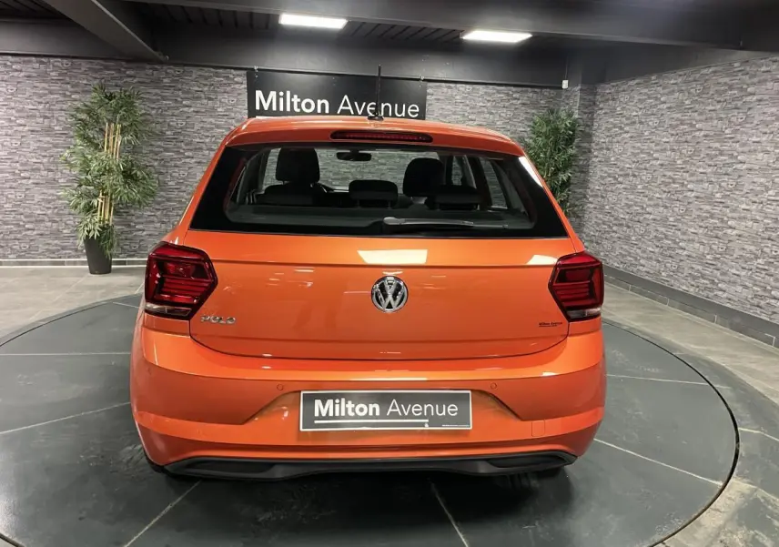 Vue arrière d'une Volkswagen Polo orange vif 2019, avec logo VW centré et feux arrière sombres en intérieur showroom.