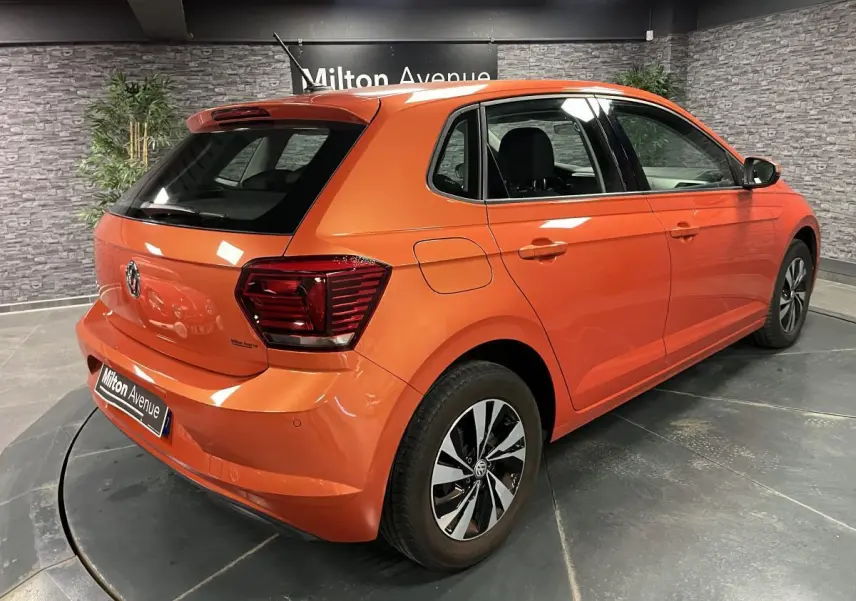 Volkswagen Polo orange vue 3/4 arrière droit, avec jantes alliage et feux arrière foncés en intérieur showroom.