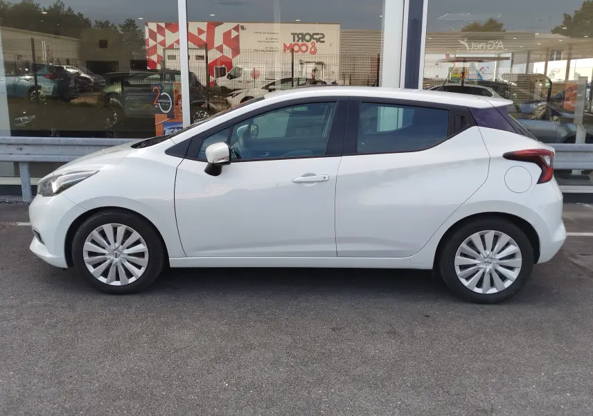 Profil côté gauche d'une Nissan Micra blanche 2019 garée devant une vitrine, avec jantes alliage et vitres teintées.