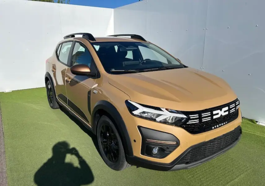 Vue 3/4 avant d'une Dacia Sandero Stepway beige Safari avec calandre noire et jantes noires sur sol vert.
