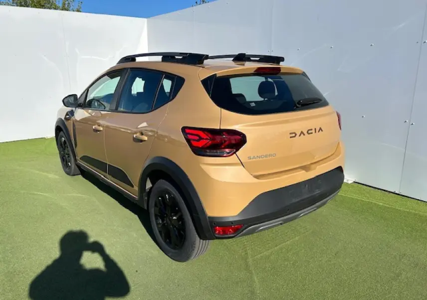 Vue 3/4 arrière droite d'une Dacia Sandero beige Safari avec barres de toit noires et jantes noires.