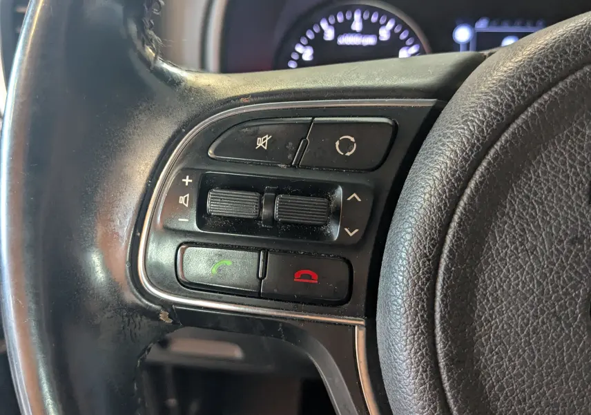Gros plan sur les commandes gauche du volant en cuir noir du Kia Sportage gris, avec boutons de volume et téléphone.