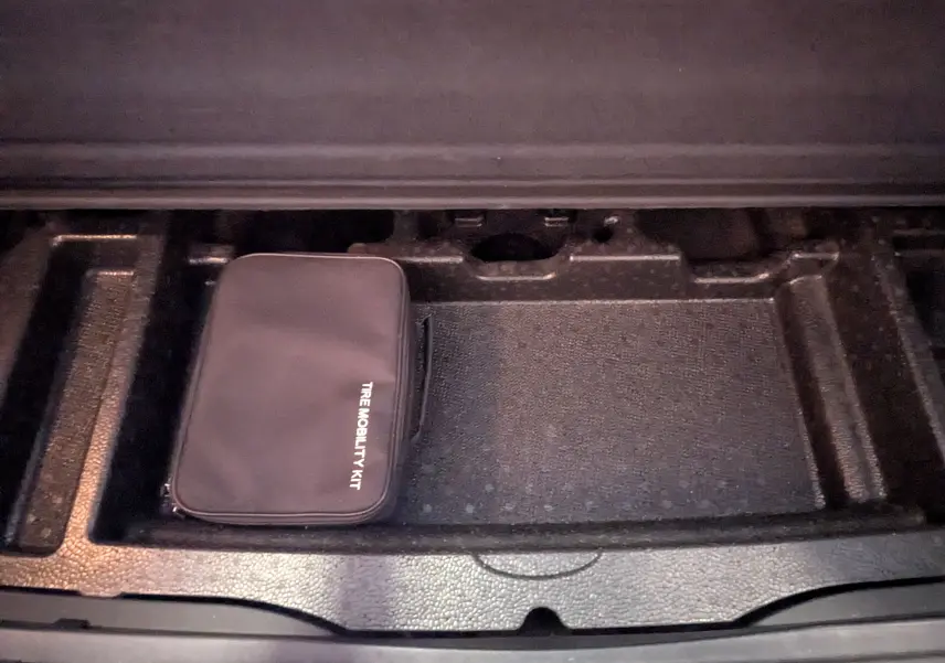 Compartiment de rangement noir dans le coffre d'un KIA Sportage gris, contenant un kit de mobilité pneus.