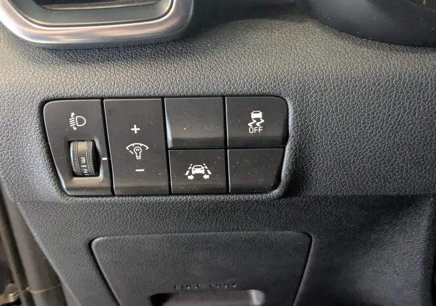 Gros plan sur les commandes noires du tableau de bord côté gauche du Kia Sportage gris, avec réglage phares et aides à la conduite.