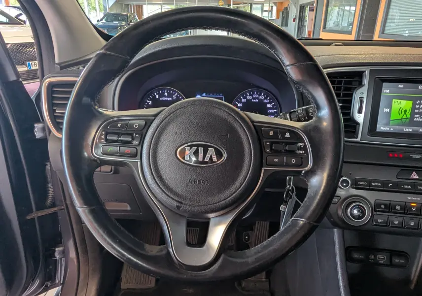 Vue intérieure centrée sur le volant noir du Kia Sportage 2016 avec tableau de bord et écran multimédia visibles.