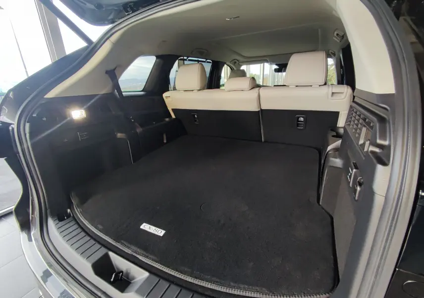 Vue arrière ouverte du coffre spacieux du Mazda CX-80 gris foncé avec banquette arrière beige visible.