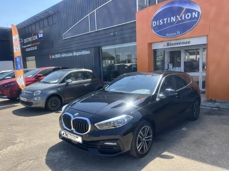 BMW Série 1 noire vue en 3/4 avant droit, garée devant un concessionnaire Distinxion avec calandre chromée visible.
