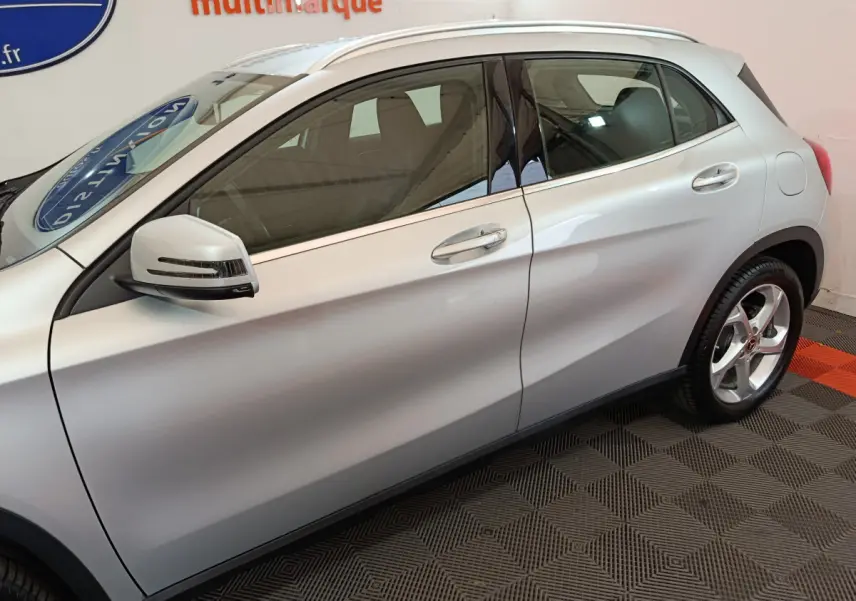 Profil côté gauche d'un Mercedes GLA 200 d gris clair avec rétroviseur et jante alliage visibles en intérieur showroom.