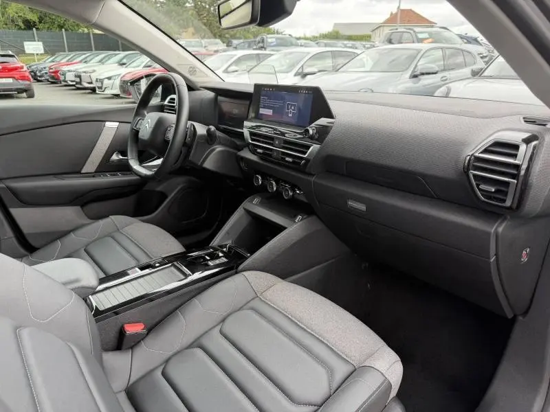 Intérieur de la Citroën C4 X 2024 vu côté conducteur, avec tableau de bord moderne et sièges gris en tissu et cuir.
