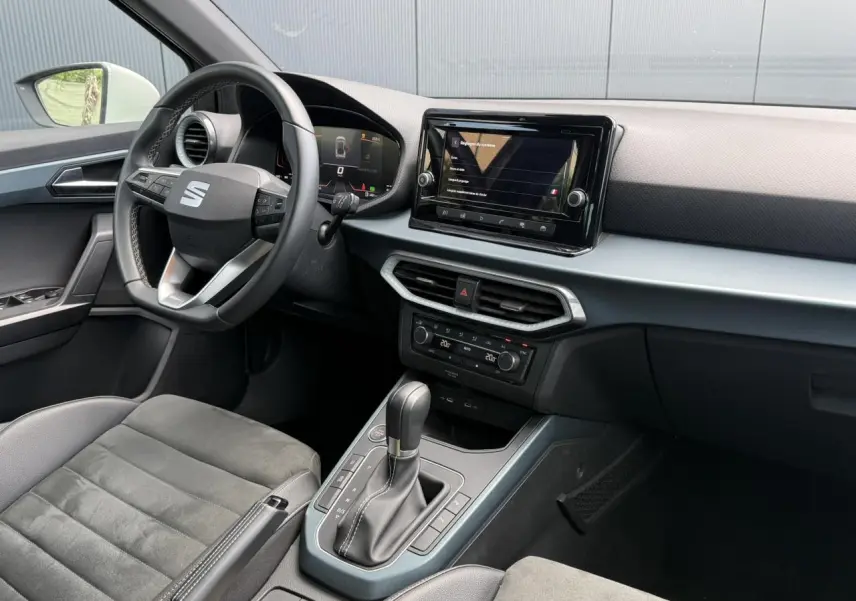 Vue intérieure côté conducteur du SEAT Arona 2025, mettant en valeur le volant, l'écran tactile et la sellerie en suédine noire.