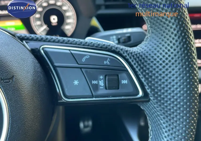 Gros plan sur les commandes gauche du volant perforé d’une Audi A3 Sportback noire, avec tableau de bord numérique en arrière-plan.