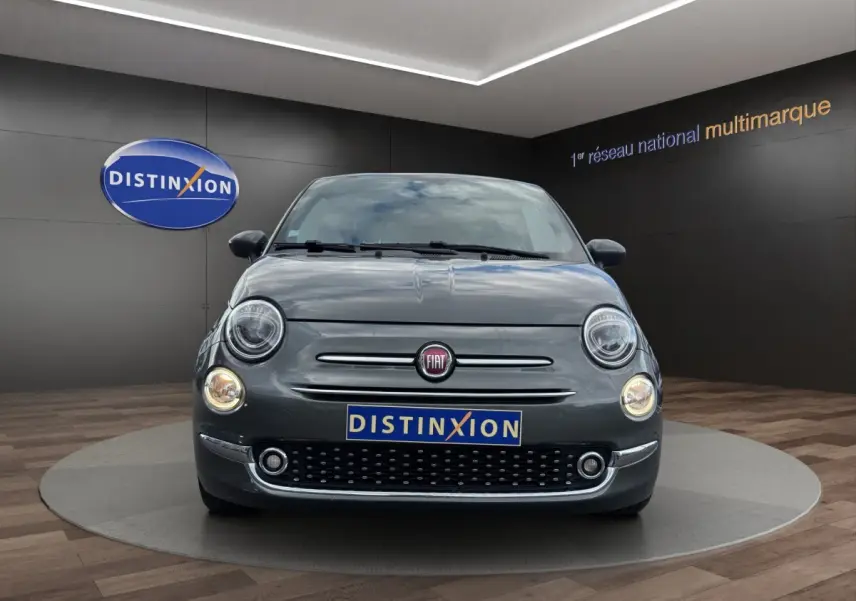 Vue avant de la Fiat 500 gris foncé avec phares ronds allumés et calandre chromée dans un showroom.