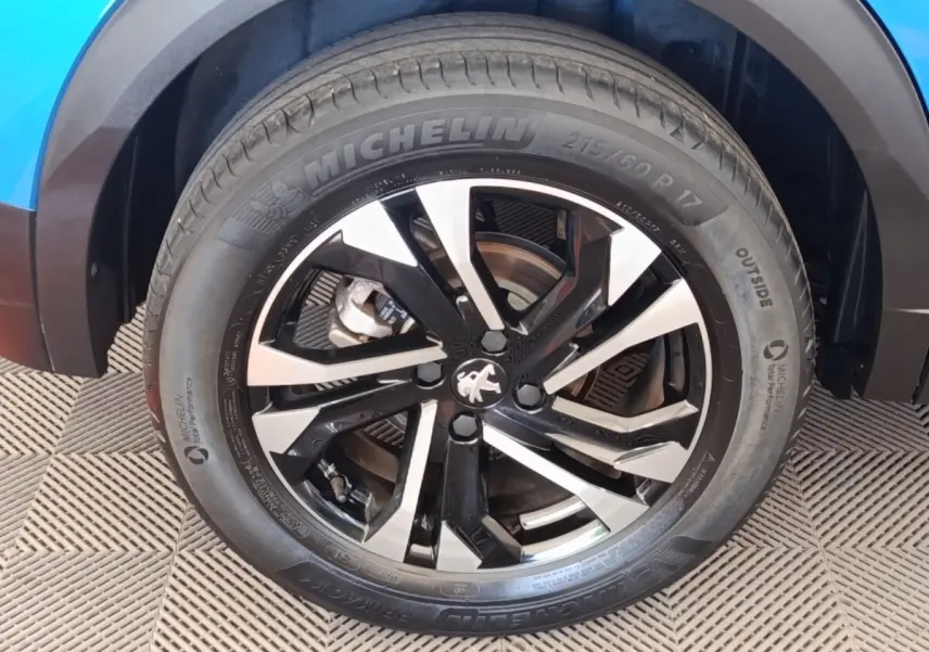 Gros plan sur la roue avant gauche du Peugeot 2008 bleu, mettant en valeur la jante alliage noire et argentée avec logo Peugeot.