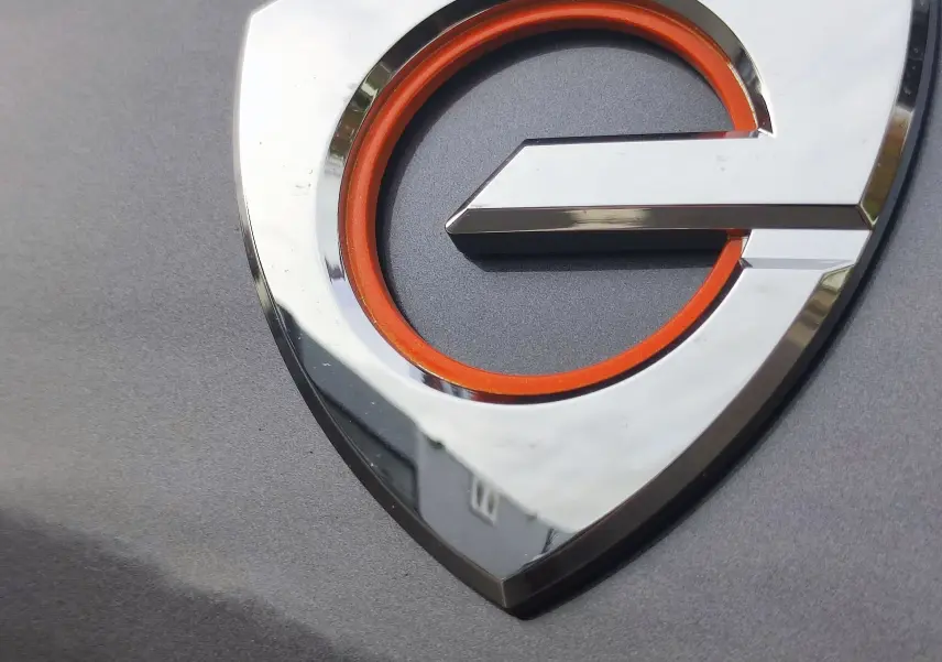 Gros plan sur le logo e hybride rechargeable orange et chrome sur la carrosserie gris foncé du Mazda MX-30 2023.
