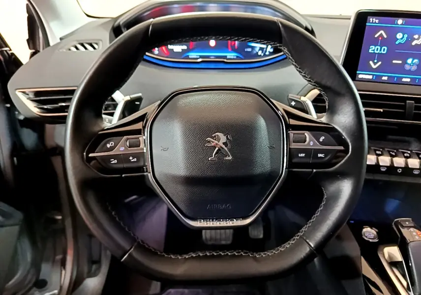 Volant cuir noir avec logo Peugeot, tableau de bord digital et écran tactile de climatisation dans un 5008 gris clair, vue intérieure frontale.
