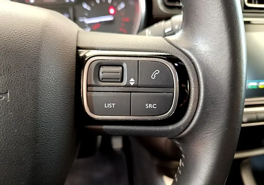 Gros plan sur les commandes gauche du volant cuir noir d'une Citroën C3 gris clair, avec tableau de bord flou en arrière-plan.