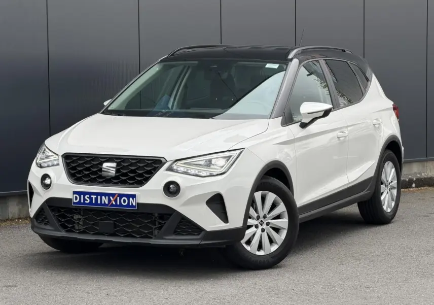 Vue 3/4 avant droite d'un SEAT Arona blanc avec toit noir et jantes alliage, stationné devant un mur gris.