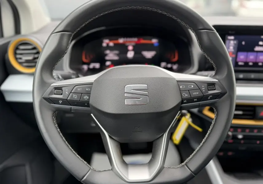 Vue rapprochée du volant noir du SEAT Arona 2025 avec commandes intégrées et tableau de bord digital flou en arrière-plan.