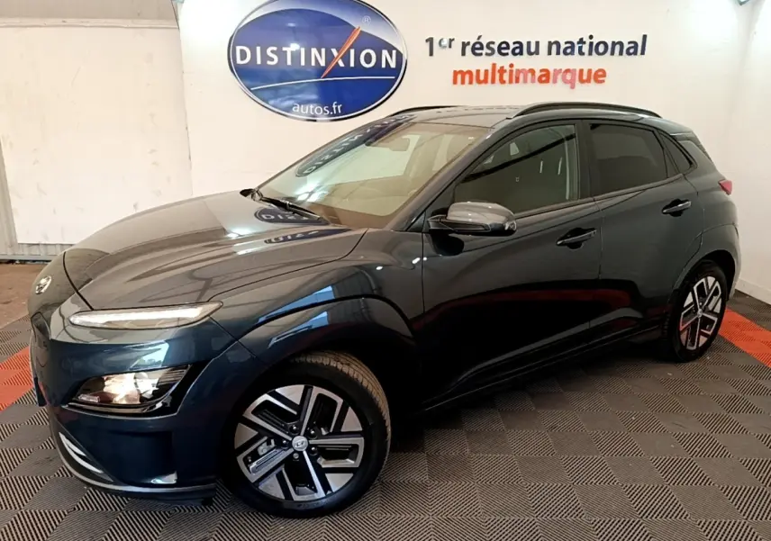 Hyundai Kona électrique bleu en 3/4 avant droit, avec jantes alliage et rétroviseurs chromés dans un showroom.