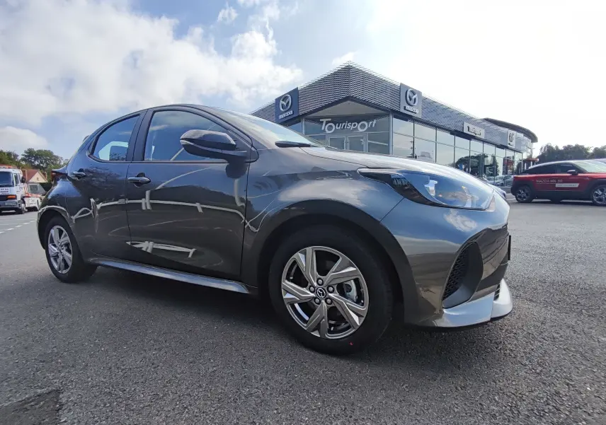 Vue 3/4 avant côté gauche d'une Mazda2 gris foncé hybride stationnée devant un concessionnaire Mazda.