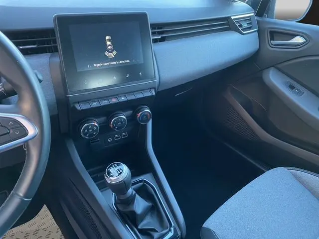 Vue intérieure côté passager de la Renault Clio V PH2 TCE 90 Evolution 2024, tableau de bord gris avec écran tactile et levier de vitesse manuel.