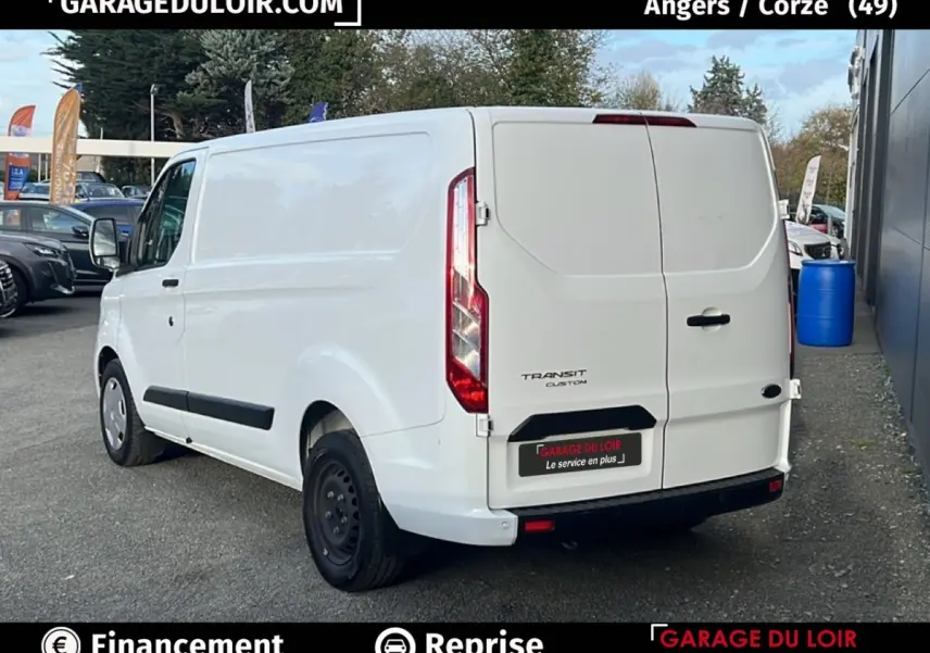 Vue 3/4 arrière droite d'un Ford Transit Custom fourgon blanc avec portes arrière fermées sur parking extérieur.