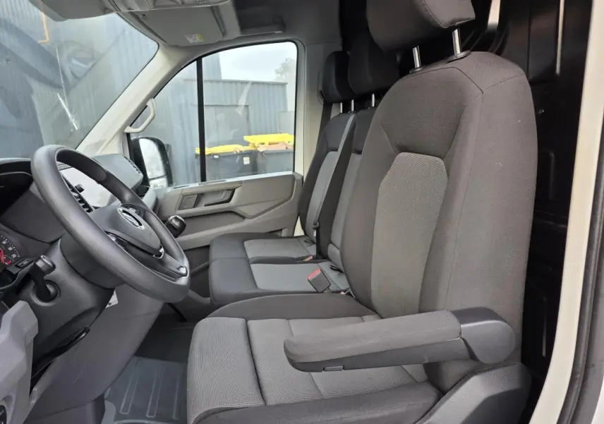Vue intérieure côté conducteur du Volkswagen Crafter blanc 2023, sièges tissu gris avec accoudoir et volant multifonctions.