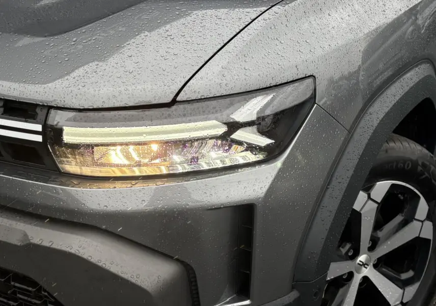 Gros plan sur l'avant droit gris schiste du Dacia Duster 2026 avec phare allumé et jante alliage diamantée.