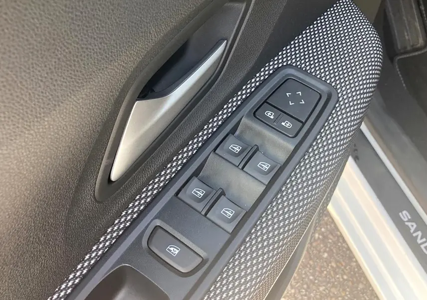 Gros plan sur la commande des vitres électriques côté conducteur avec poignée argentée, intérieur tissu gris et seuil de porte blanc Sandero.