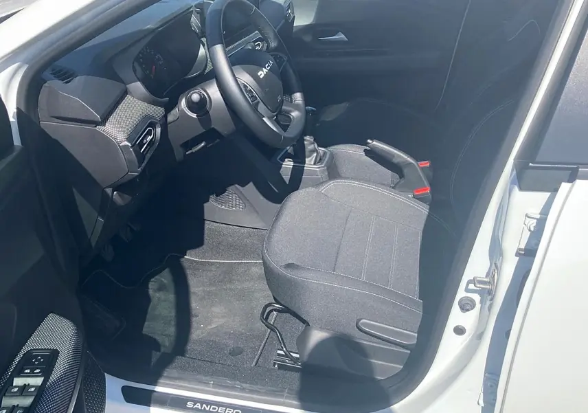 Vue intérieure côté conducteur de la Dacia Sandero blanche 2025, volant, siège tissu gris et levier de vitesse manuel visibles.