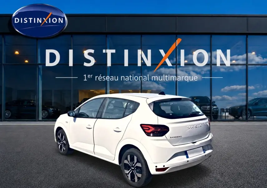 Vue 3/4 arrière droite d'une Dacia Sandero blanche 2025 avec jantes alu et feux arrière LED, devant un showroom Distinxion.