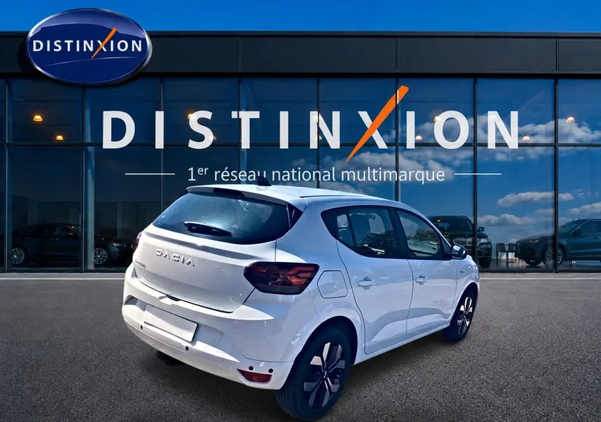 Vue 3/4 arrière droite d'une Dacia Sandero blanche 2025 avec jantes alu et becquet arrière, devant un showroom Distinxion.