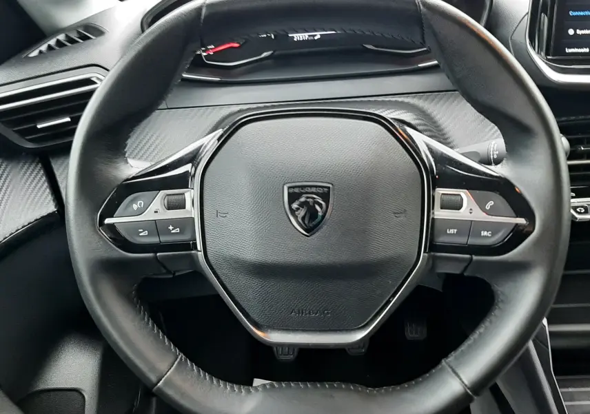 Vue rapprochée du volant noir de la Peugeot 208 2024, avec tableau de bord numérique et détails en carbone gris.