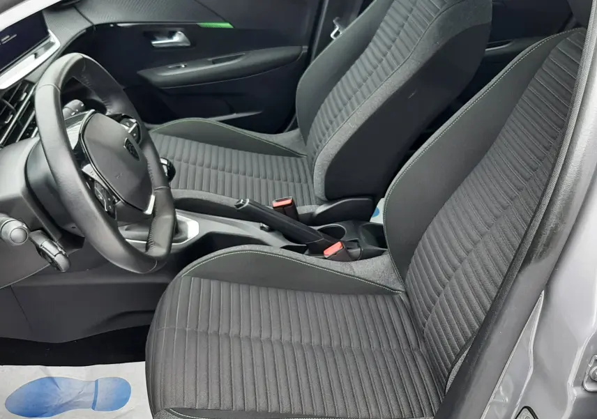Vue intérieure côté conducteur de la Peugeot 208 2024 gris selenium, sièges tissu gris avec coutures vertes et volant multifonction.
